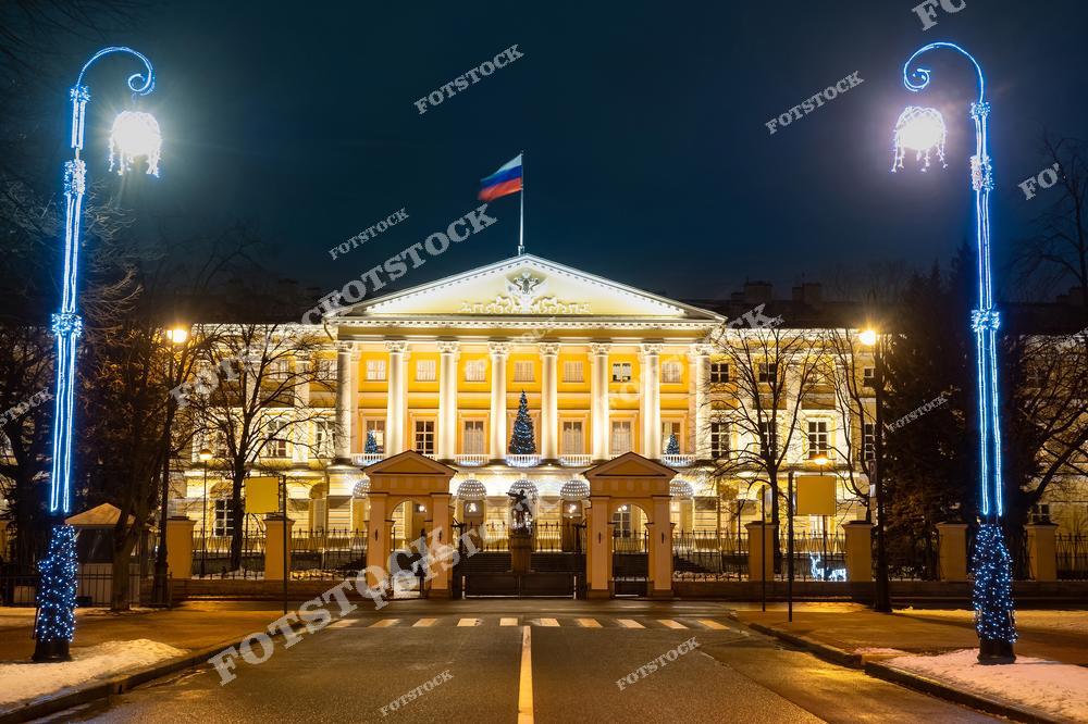 На фотографии изображён Смольный дворец, сияющий в праздничной подсветке в зимний вечер. Украшенные гирляндами фонари на переднем плане подчёркивают торжественную атмосферу. На фасаде дворца видна новогодняя ёлка, добавляющая праздничного настроения. Этот исторический памятник, символ управления и культуры Санкт-Петербурга, выглядит особенно впечатляюще ночью.